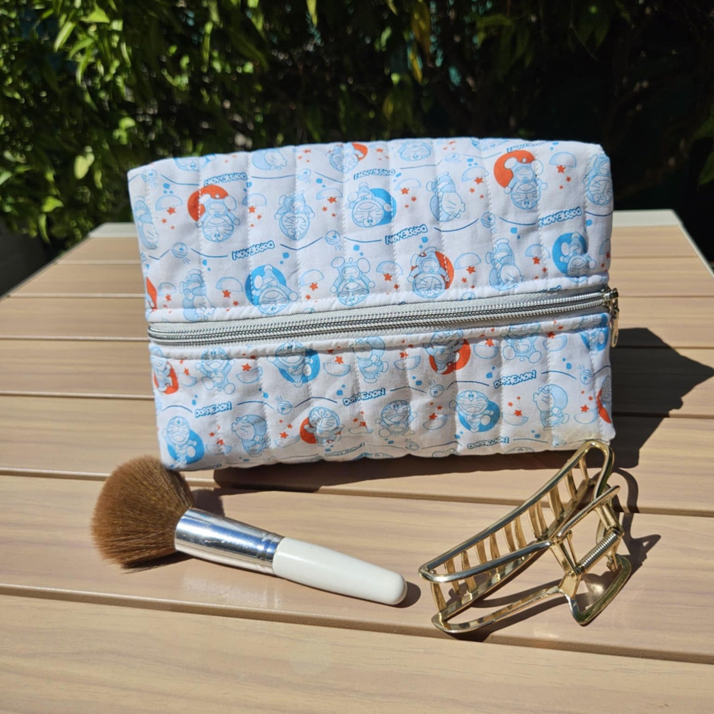 Makeup bag with a pattern, brush, and eyelash curler on a wooden surface with greenery in the background