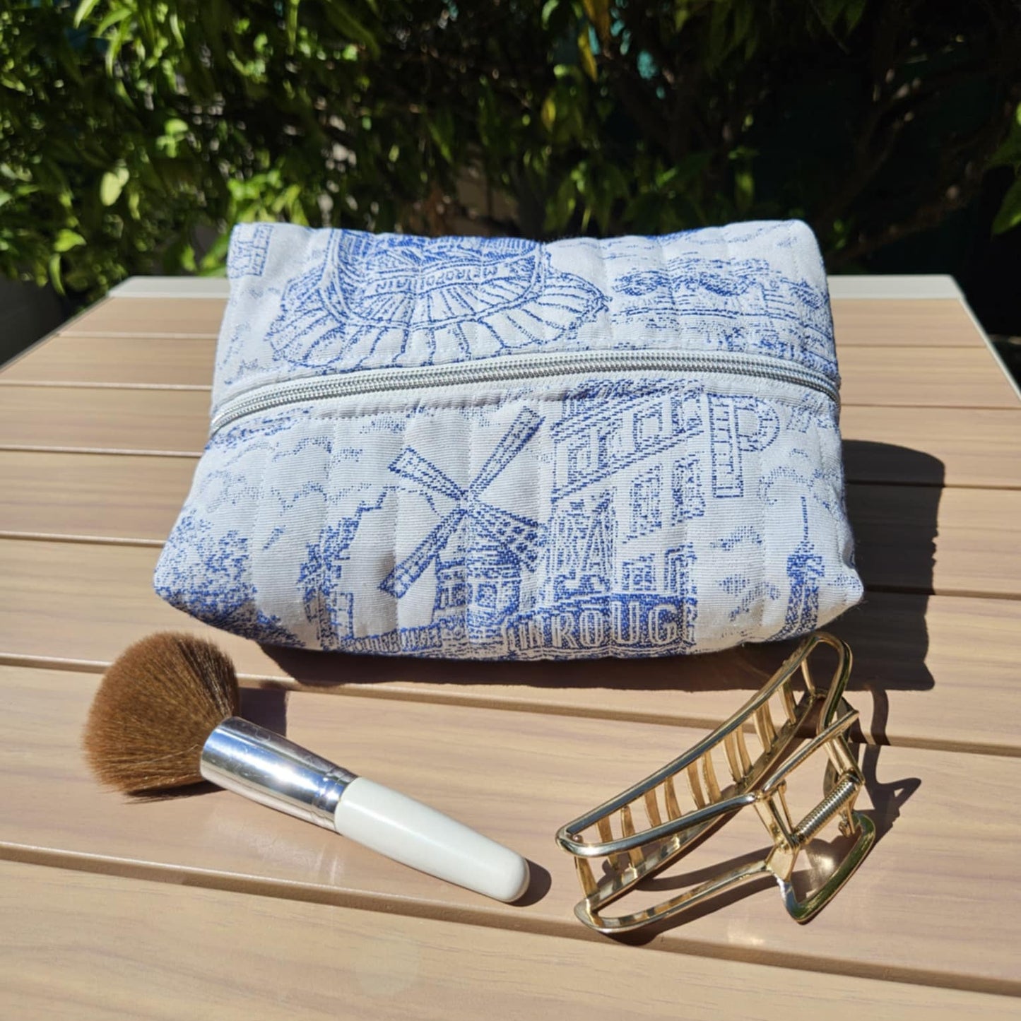 Blue and white patterned pouch with a makeup brush and gold clip on a wooden surface with greenery in the background