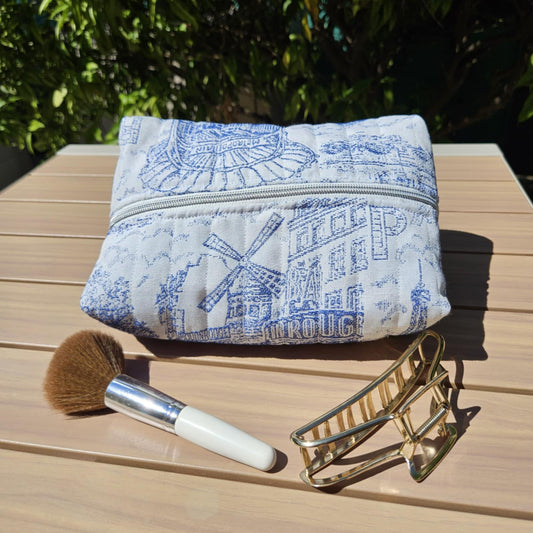 Blue and white patterned pouch with a makeup brush and gold clip on a wooden surface with greenery in the background
