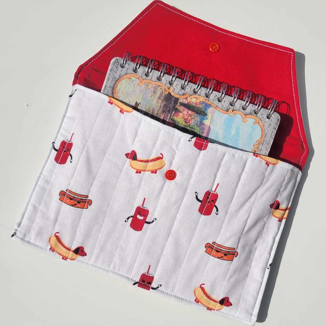 Handmade, lightly quilted notebook holder with a snap closure. Exterior fabric is a playful Dachshund hot dog and cola pattern with a bright red interior fabric.